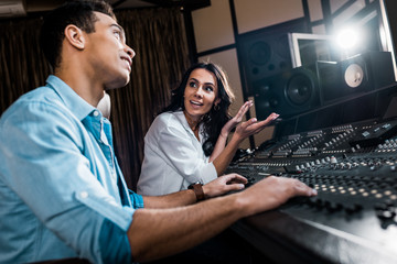 Fototapeta na wymiar selective focus of pretty sound producer gesturing near mixed raced colleague working at mixing console