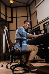 Fototapeta na wymiar smiling mixed race sound producer sitting in office chair in recording studio and looking at camera
