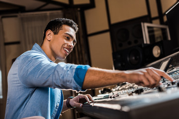 selective focus of smiling mixed race sound producer working at mixing console in recording studio