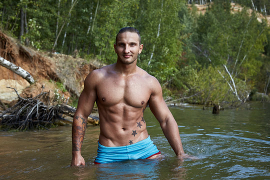 Male sexy fit model looks into the camera standing waist - deep in the water against the background of a wild forest, sandy beach and clear water