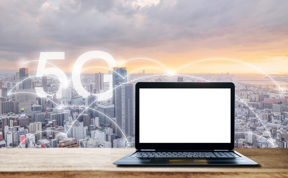 Internet networking, 5G and wireless technology. Computer laptop with blank white screen, on wooden desk and city view in sunset with 5G internet networking