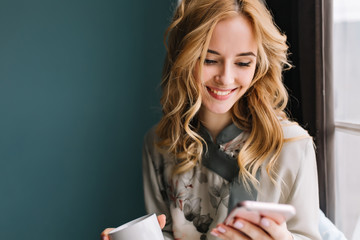 Closeup portrait of young blonde woman with wavy hair texting on phone while drinking coffee or...