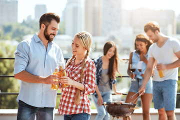 Celebrating summer. Two happy young friends clinking glasses with beer and smiling while standing on the roof with friends. Barbecue concept.