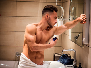 Handsome shirtless athletic young man in his home bathroom, spraying deodorant on armpits
