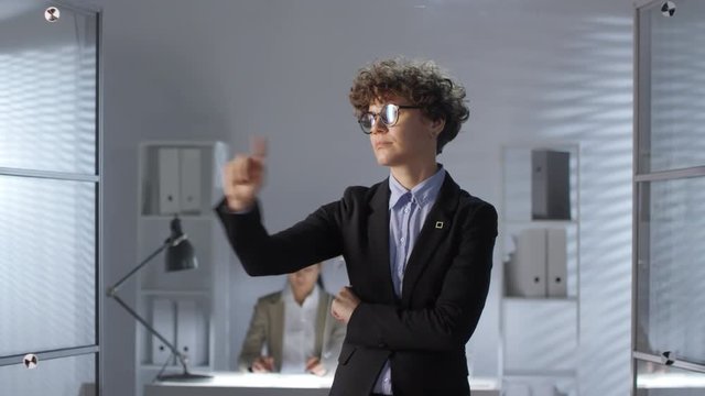 Businesswoman in formal suit tapping and swiping on invisible augmented reality screen while her female colleague working on computer at desk in office. Footage suitable for adding AR elements