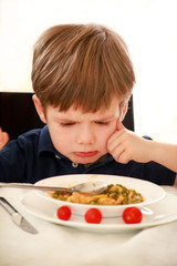 Hungry child sitting in chair at table in kitchen and despises, disapproves of hot meal, because he does not like lunch. Kid is having of cooked peas with tomato for lunch. Healthy diet food concept.