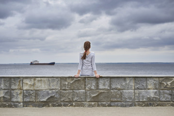 Rear view of a girl sitting on the embankment of the river. A ship is sailing on the horizon. There are heavy clouds in the sky.