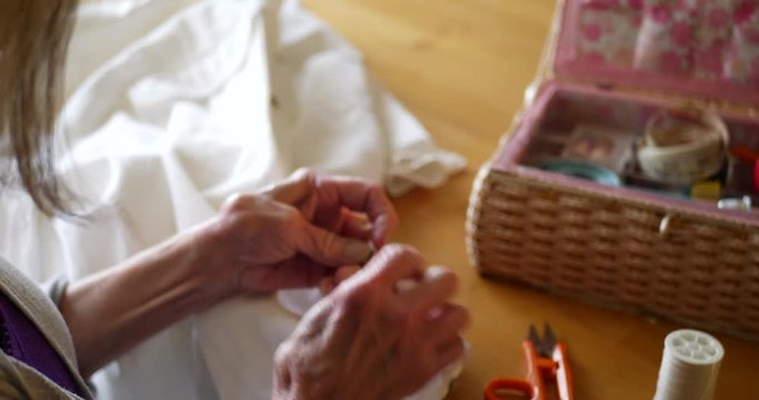 Close up on the hands of an old woman cutting the thread of an old button on a white dress shirt to replace it with a new one with her sewing kit.