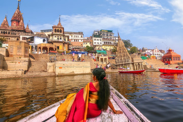 Female tourist in traditional dress (saree) on a wooden boat overlooking the historic Varanasi city architecture and Ganges river ghat at Uttar Pradesh India