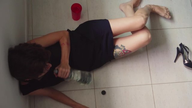 high angle of a boozy woman trying to get up of the floor while drinking from a bottle of vodka