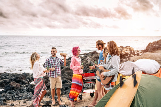 Happy surfer friends making party at sunset while camping next the beach - Young people having fun and drinking beer outdoor - Millennial, summer, vacation and youth holidays lifestyle concept