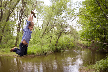 Cute boy rides a homemade bungee in the park. Bungee tied to a tree branch on the river bank. A...