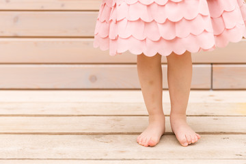 Beautifully beautiful legs of a child girl stand on a wooden terrace at home. Little girl in a pink fashionable dress barefoot on the street on a summer day. Outdoor party