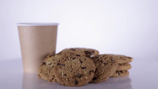 Middle shot of hot drink in a paper cup rotating with several oatmeal cookies with chocolate. Coffee or tea with oatmeal cookies for breakfast on white table and white background