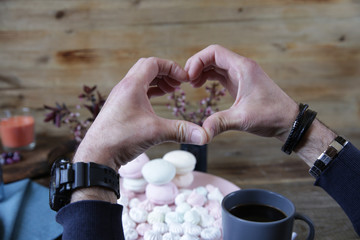 men's hands in watches and bracelets folded in the shape of a heart against the background of pink marshmallow, a cup of coffee and spring flowers on a wooden rustic background