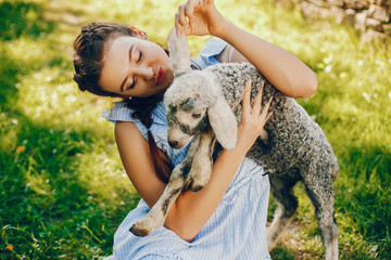 beautiful and cute girl in blue dresses with beautiful hairstyles and make-up sitting in a sunny green garden and playing with a goat