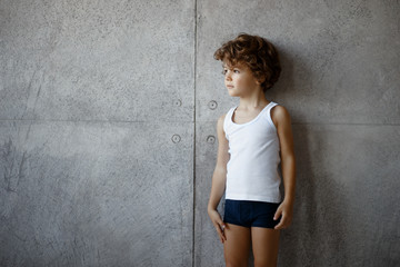 Curly cute little boy in underwear poses on concrete wall, lokking a one side, copy space.