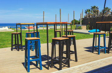 High table and chairs on wooden floor in exterior cafe restaurant with the beach and puffs