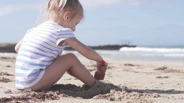 Cute little girl sitting on the sandy beach by the sea. The child buries its feet with sand. She diligently plays her game