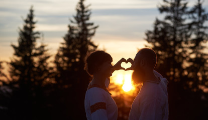 Silhouettes of happy couple in love in the evening. Bearded man and smiling woman gesturing heart shaped hands, looking at each. Beautiful sunset, cloudy sky and forest on blurred background