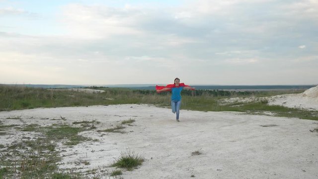 young girl dreams of becoming superhero. happy girl superhero runs across field in red cloak in front of blue sky and smiles, slow motion. happy teen girl goes in red cloak expression of dreams.