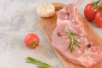 A raw piece of pork lies on a wooden cutting board. Near tomatoes, garlic and rosemary. Preparing steak for cooking.