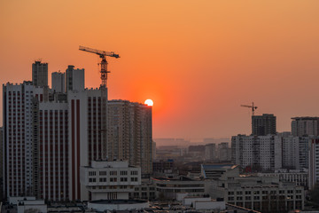 Fototapeta na wymiar Sunset sun hiding behind city's high-rise buildings, orange cloudless sky cityscape
