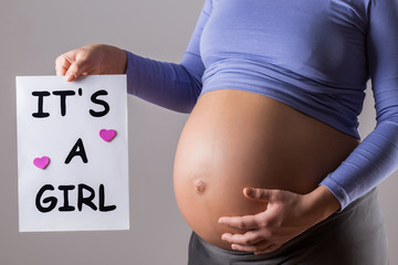 Image of close up stomach of pregnant woman holding paper with text it's a girl on gray background.