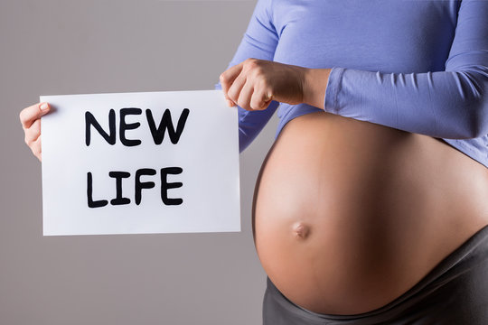 Image of close up stomach of pregnant woman holding paper with text new life on gray background.