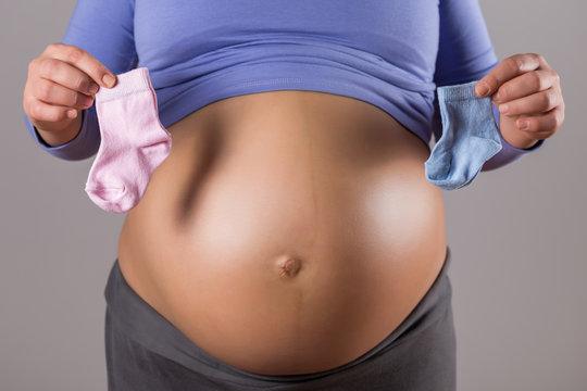 Image of stomach of pregnant woman holding pink and blue baby socks  on gray background.