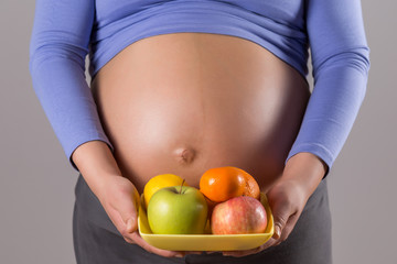 Image of close up stomach of pregnant woman holding fruit on gray background.