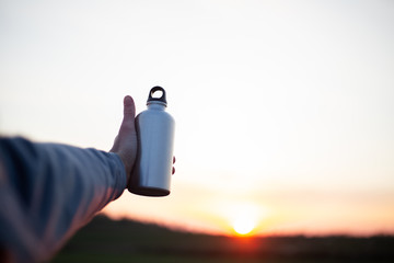 Hand of man holding aluminium bottle for water, on background of sunset.