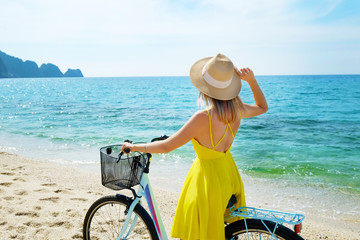 Fototapeta na wymiar Young carefree woman in bright yellow dress with bicycle at ocean beach. Unrecognizable female wearing broad brim straw hat biking on sandy sea shore on sunny day. Copy space, background.
