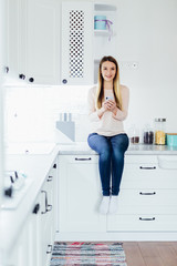 Fototapeta na wymiar Young blonde woman using mobile phone sitting in modern kitchen