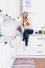 Adorable nice charming cheerful beautiful trendy stylish girl chatting on mobile sitting on table countertop in modern light white kitchen