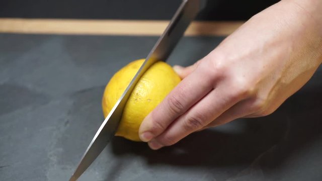 Close up, a woman's hands take a knife and cut the yellow lemon exactly in half, then take the two halves and bring it to the video camera, then lay it on the kitchen stone board with a wooden frame
