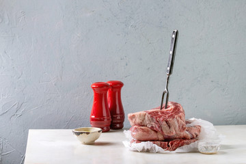 Three raw uncooked beef steaks in stack on meat fork on crumpled paper, salt, pepper and olive oil for cooking on white marble table.