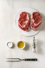 Three raw uncooked beef steaks on ceramic board with meat fork, pepper, salt and olive oil in row over white marble background. Flat lay, space