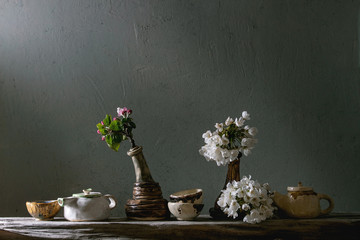 Variety of craft handmade ceramic teapots and cups for tea ceremony standing with spring blossom branches in vase on old wooden shelf in dark room.