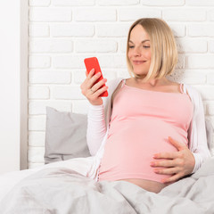 Happy smiling pregnant woman lies in bed and looks at mobile phone