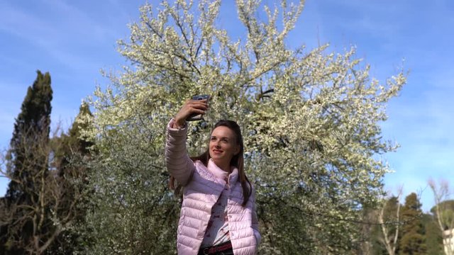 Pretty girl with long brown hair and adorable smile making selfie photos on smartphone on the background of beautiful blooming trees. Woman enjoying springtime in the park
