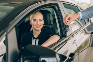 woman sitting in new car while salesman holding car keys beside her in car dealership showroom