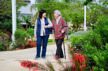 Cheerful female carer and an older woman with a walking stick stroll in a city park. Both are in a...