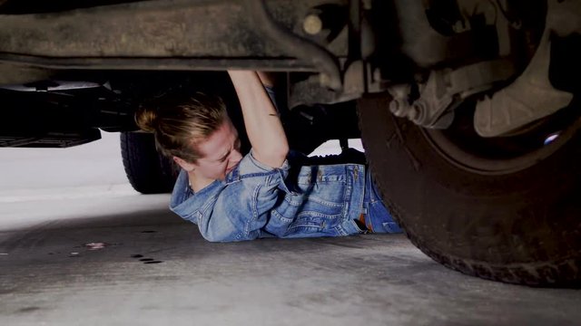 A woman on the ground underneath a vehicle, having troubles while going through routine maintenance, 23.98 fps.