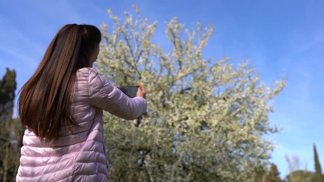 Beautiful young girl with long brown hair and adorable face dressed in pink jacket taking photos of fresh blooming trees with smartphone