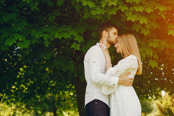 Fototapeta premium A beautiful and gentle girl with light hair and a white dress is walking in a sunny summer forest with her handsome guy in a white shirt and dark pants