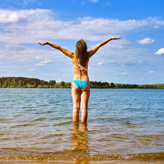 Teenager girl wearing bikini with spreading hands jumps high above the water of a beautiful lake enjoying warm weather at summer vacations. Happy summer vacations.