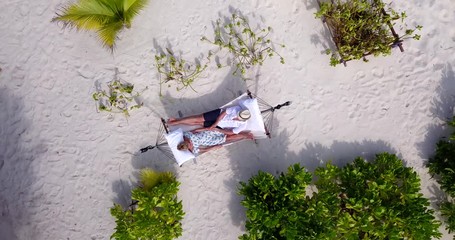 Top view, couple relaxing in hammock together on secluded sandy beach, slow aerial rise rotating shot