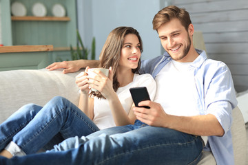 Young couple watching online content in a smart phone sitting on a sofa at home in the living room.