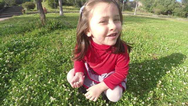 4k Little girl at park makes cheerful expression, happy child girl playing on grass in spring picking flowers